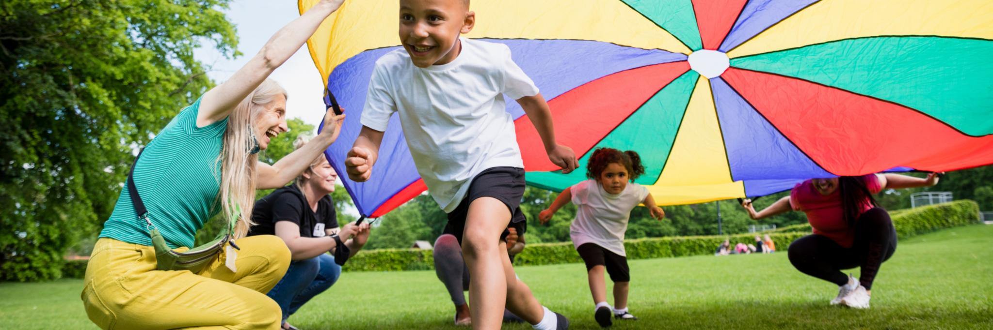 Man sieht fröhliche Kinder, die unter einem bunten großen Tuch, das von Erwachsenen gehalten wird, über die Wiese rennen.